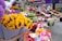 Bright and colorful sunflowers are prominently displayed alongside an assortment of other flowers like gerberas and chrysanthemums. The background reveals a bustling street flower market with a variety of floral arrangements, set against the facade of a pharmacy.