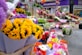 Bright and colorful sunflowers are prominently displayed alongside an assortment of other flowers like gerberas and chrysanthemums. The background reveals a bustling street flower market with a variety of floral arrangements, set against the facade of a pharmacy.