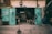 A weathered garage with teal double doors wide open, revealing a cluttered workshop inside. Various tools and equipment are visible, with wires and hoses hanging from the doors. A small chair sits in the center, and a large blue barrel stands to the side. The ambiance suggests a well-used and functional workspace.
