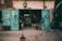 A weathered garage with teal double doors wide open, revealing a cluttered workshop inside. Various tools and equipment are visible, with wires and hoses hanging from the doors. A small chair sits in the center, and a large blue barrel stands to the side. The ambiance suggests a well-used and functional workspace.