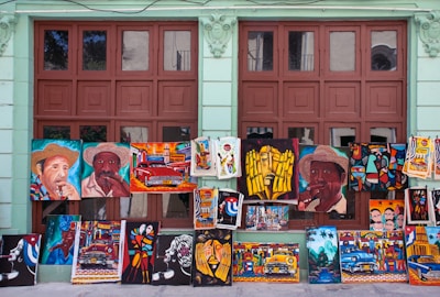 A variety of colorful paintings displayed against a wall with large windows. The paintings depict various subjects including portraits of people wearing hats, vintage cars, abstract art, and cultural scenes. The background wall is painted light green and the windows have a brown frame.
