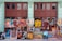 A variety of colorful paintings displayed against a wall with large windows. The paintings depict various subjects including portraits of people wearing hats, vintage cars, abstract art, and cultural scenes. The background wall is painted light green and the windows have a brown frame.