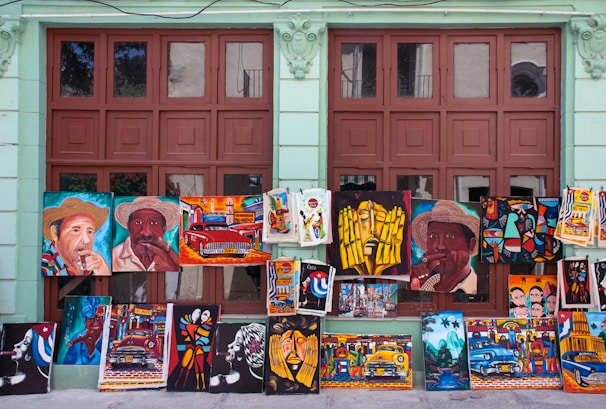 A variety of colorful paintings displayed against a wall with large windows. The paintings depict various subjects including portraits of people wearing hats, vintage cars, abstract art, and cultural scenes. The background wall is painted light green and the windows have a brown frame.