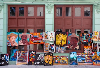 A variety of colorful paintings displayed against a wall with large windows. The paintings depict various subjects including portraits of people wearing hats, vintage cars, abstract art, and cultural scenes. The background wall is painted light green and the windows have a brown frame.