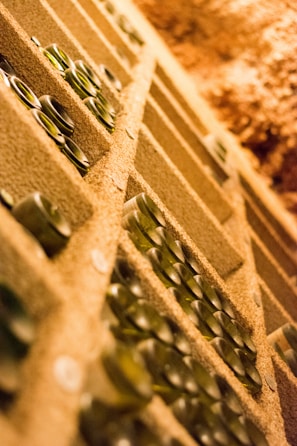 Rows of wine bottles are stored horizontally in a cellar, with the focus on the orderly arrangement of the bottles in a dimly lit, rustic environment. The angle gives a unique perspective, emphasizing depth and the texture of the shelving.
