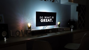 A modern desk setup with a computer monitor displaying the text 'DO WHAT IS GREAT.' Two stylish light bulbs are positioned symmetrically on either side of the monitor, adding a warm glow to the scene. The desk is equipped with a white keyboard and mouse, and features the word 'LOVE' in large letters. Speakers and other tech gadgets are neatly arranged around the setup.