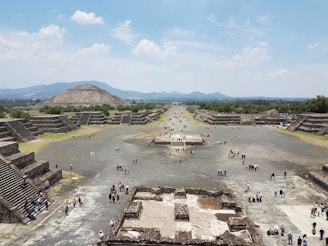 A vast archaeological site with large, ancient stone pyramids and stepped platforms, surrounded by crowds of visitors walking along a wide, open pathway. The landscape includes distant mountains under a partly cloudy sky.