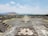 A vast archaeological site with large, ancient stone pyramids and stepped platforms, surrounded by crowds of visitors walking along a wide, open pathway. The landscape includes distant mountains under a partly cloudy sky.