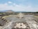 A vast archaeological site with large, ancient stone pyramids and stepped platforms, surrounded by crowds of visitors walking along a wide, open pathway. The landscape includes distant mountains under a partly cloudy sky.