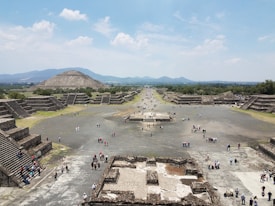 A vast archaeological site with large, ancient stone pyramids and stepped platforms, surrounded by crowds of visitors walking along a wide, open pathway. The landscape includes distant mountains under a partly cloudy sky.