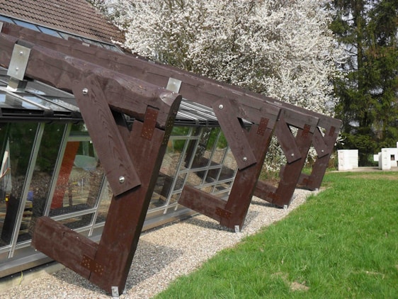 Large wooden structures with angular designs are placed along a glass window wall. The wooden beams are dark brown and are secured with metal brackets. Behind the structures, a tree full of white blossoms is visible, adding a natural contrast to the scene. The foreground features a grassy area with a hint of gravel visible near the base of the structures.