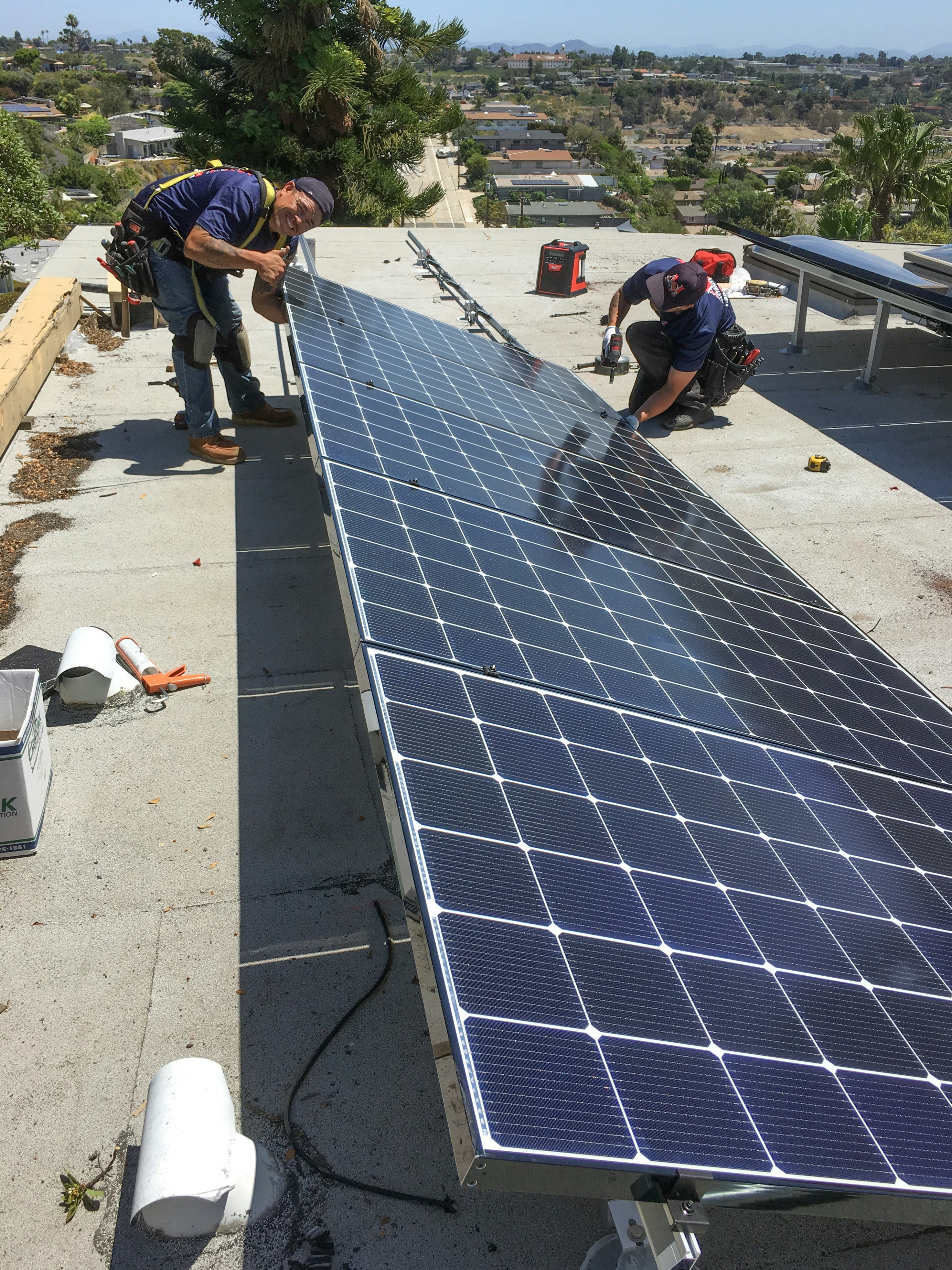 Two men are working on installing large solar panels on a rooftop. One is kneeling and appears to be securing the panel with tools, while the other is standing and adjusting the panel. The background shows a view of a suburban area with houses and trees under a clear blue sky.