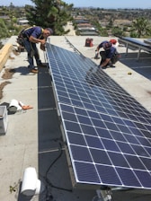 Two men are working on installing large solar panels on a rooftop. One is kneeling and appears to be securing the panel with tools, while the other is standing and adjusting the panel. The background shows a view of a suburban area with houses and trees under a clear blue sky.