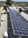 Two men are working on installing large solar panels on a rooftop. One is kneeling and appears to be securing the panel with tools, while the other is standing and adjusting the panel. The background shows a view of a suburban area with houses and trees under a clear blue sky.