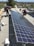 Two men are working on installing large solar panels on a rooftop. One is kneeling and appears to be securing the panel with tools, while the other is standing and adjusting the panel. The background shows a view of a suburban area with houses and trees under a clear blue sky.