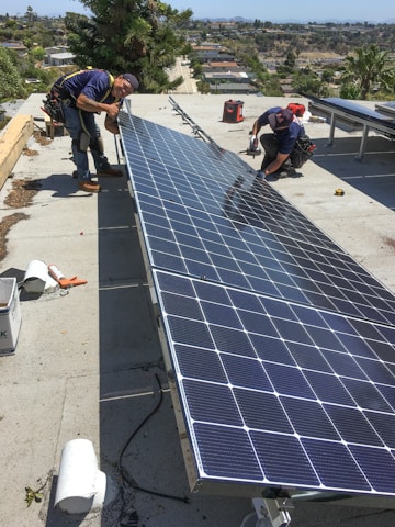 Two men are working on installing large solar panels on a rooftop. One is kneeling and appears to be securing the panel with tools, while the other is standing and adjusting the panel. The background shows a view of a suburban area with houses and trees under a clear blue sky.