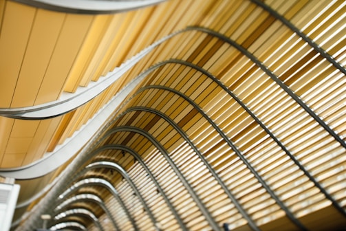 Curved architectural design featuring a series of parallel, wavy lines and metallic structures in shades of yellow and beige.