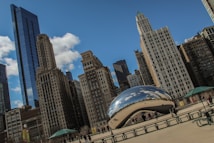 A cityscape featuring a large, reflective sculpture known as Cloud Gate in a public plaza with tall skyscrapers in the background. The sky is blue with a few clouds, and people are walking around, some taking photos.