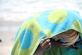 A child is wrapped in a towel with large blue and green circles, standing on a beach. The head is slightly bowed, and the sandy hand is gripping the towel. The blurred background suggests a seashore with gentle waves.