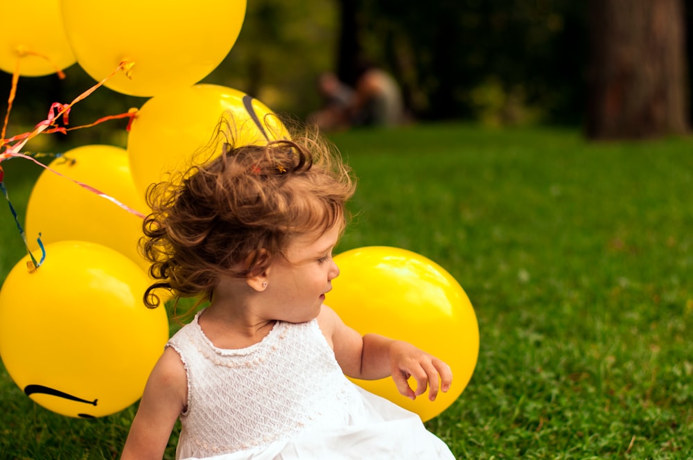 A little girl in a white dress sitting down next to a bunch of yellow balloons.