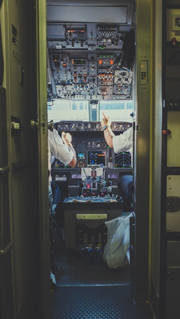 View into an airplane cockpit from a doorway, showing two pilots interacting with the control panels. The cockpit is filled with numerous dials, switches, and displays, creating a complex and technical environment. The pilots are in white uniforms, engaged in conversation or conducting checks.