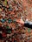 A hand is placing a piece of chewing gum onto a wall covered in numerous pieces of colorful gum. The wall has an assortment of colors and textures, creating a complex and vibrant pattern.