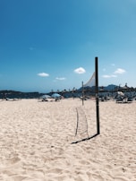 A sunny beach scene with a volleyball net stretching across the sandy area in the foreground. In the background, there are several beach umbrellas with people lounging beneath them. The sky is clear with a few small clouds scattered, and the sea is visible in the distance.