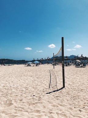 A sunny beach scene with a volleyball net stretching across the sandy area in the foreground. In the background, there are several beach umbrellas with people lounging beneath them. The sky is clear with a few small clouds scattered, and the sea is visible in the distance.