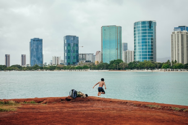 A young person is mid-jump on a red dirt path next to a body of water, with a large backpack nearby. In the background, a cityscape with modern high-rise buildings towers over a line of trees.