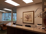 An office reception area featuring a wooden counter, a comfortable chair, and a corner with a desk and office supplies. The walls are decorated in neutral tones, with a framed artwork depicting a floral arrangement. Large windows reveal an urban exterior with trees and buildings visible.