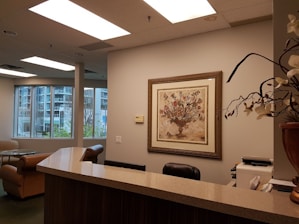 An office reception area featuring a wooden counter, a comfortable chair, and a corner with a desk and office supplies. The walls are decorated in neutral tones, with a framed artwork depicting a floral arrangement. Large windows reveal an urban exterior with trees and buildings visible.
