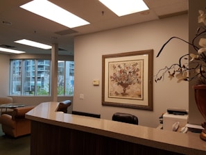 An office reception area featuring a wooden counter, a comfortable chair, and a corner with a desk and office supplies. The walls are decorated in neutral tones, with a framed artwork depicting a floral arrangement. Large windows reveal an urban exterior with trees and buildings visible.
