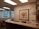 An office reception area featuring a wooden counter, a comfortable chair, and a corner with a desk and office supplies. The walls are decorated in neutral tones, with a framed artwork depicting a floral arrangement. Large windows reveal an urban exterior with trees and buildings visible.