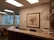 An office reception area featuring a wooden counter, a comfortable chair, and a corner with a desk and office supplies. The walls are decorated in neutral tones, with a framed artwork depicting a floral arrangement. Large windows reveal an urban exterior with trees and buildings visible.