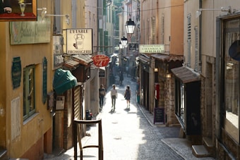 A narrow cobblestone street lined with colorful buildings featuring various shop signs. Two people walk hand in hand down the street, bathed in soft sunlight. The ambiance feels quaint and cozy, with small awnings and window shutters adding to the charm.