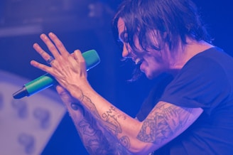 A person passionately singing into a green microphone on stage, with tattoos visible on their arm. The ambient lighting casts a blue hue over the scene, creating a dynamic and energetic atmosphere.