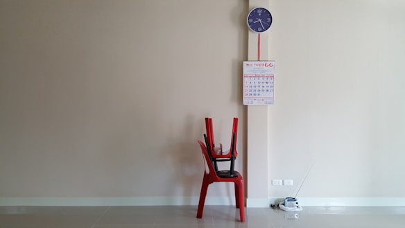 A minimalistic room setup features a red plastic chair with two other chairs stacked on top. Above the chair, a wall displays a calendar and a blue wall clock. An outlet and a small device with an antenna are placed on the floor.