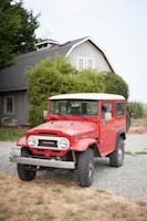 A classic red Toyota Land Cruiser parked on a gravel driveway in front of a traditional house. The house has a grey facade, white-framed windows, and is surrounded by lush greenery. The vehicle appears well-maintained with clean tires and a bright exterior.
