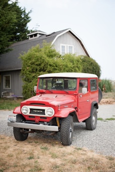 A classic red Toyota Land Cruiser parked on a gravel driveway in front of a traditional house. The house has a grey facade, white-framed windows, and is surrounded by lush greenery. The vehicle appears well-maintained with clean tires and a bright exterior.