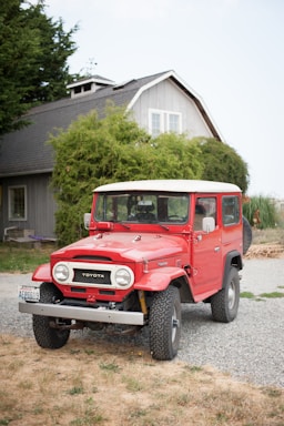 A classic red Toyota Land Cruiser parked on a gravel driveway in front of a traditional house. The house has a grey facade, white-framed windows, and is surrounded by lush greenery. The vehicle appears well-maintained with clean tires and a bright exterior.