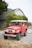 A classic red Toyota Land Cruiser parked on a gravel driveway in front of a traditional house. The house has a grey facade, white-framed windows, and is surrounded by lush greenery. The vehicle appears well-maintained with clean tires and a bright exterior.
