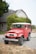 A classic red Toyota Land Cruiser parked on a gravel driveway in front of a traditional house. The house has a grey facade, white-framed windows, and is surrounded by lush greenery. The vehicle appears well-maintained with clean tires and a bright exterior.