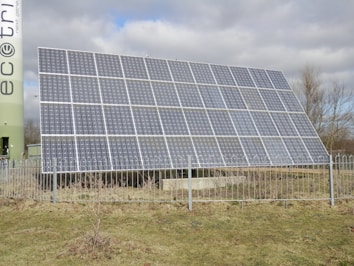 A large solar panel array is installed outdoors, surrounded by a metal fence. Behind the panels, there are leafless trees and a cloudy sky. Part of a cylindrical structure with text is visible on the left side.