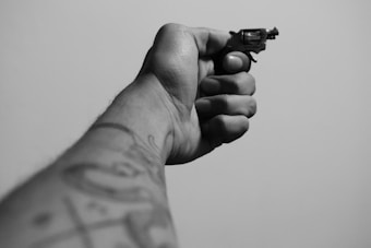 A close-up black and white photograph of a hand holding a small revolver. The arm is extended, and tattoos are visible on the arm. The focus is on the gun, while the background is a plain surface.