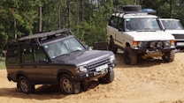 Two off-road vehicles are navigating a sandy terrain in a forested area. The first vehicle is a dark gray Range Rover equipped with roof racks and off-road tires. The second vehicle is a white Range Rover with similar off-road features, including a roof rack and sturdy tires. Both vehicles appear to be built for rugged conditions.