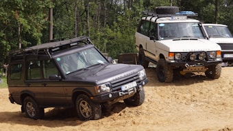 Two off-road vehicles are navigating a sandy terrain in a forested area. The first vehicle is a dark gray Range Rover equipped with roof racks and off-road tires. The second vehicle is a white Range Rover with similar off-road features, including a roof rack and sturdy tires. Both vehicles appear to be built for rugged conditions.