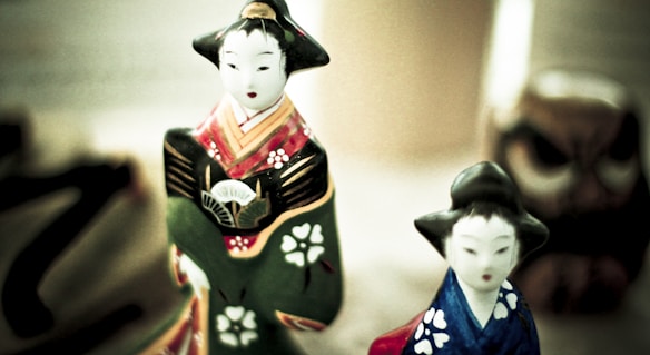 Two traditional Japanese figurines in kimonos, one in green and the other in blue. Both have delicate facial features and are adorned with intricate patterns. A blurred background suggests other cultural items.