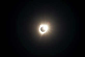 A solar eclipse is captured with the sun appearing to be almost entirely covered by the moon, creating a corona effect. Light flares can be seen radiating from the sun's edge, contrasting against the dark sky.