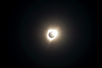 A solar eclipse is captured with the sun appearing to be almost entirely covered by the moon, creating a corona effect. Light flares can be seen radiating from the sun's edge, contrasting against the dark sky.