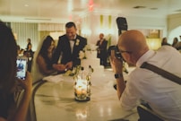 A couple, possibly newlyweds, is cutting a cake with a professional photographer taking pictures of the moment. The setting appears to be a warmly lit reception area with other guests in the background. A smartphone is visible in the foreground, capturing the event.